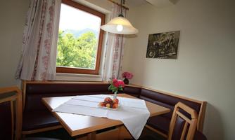 A cozy dining area with a table and a bench. A small table is decorated with fruit and a vase of flowers, and the window offers a view of greenery.