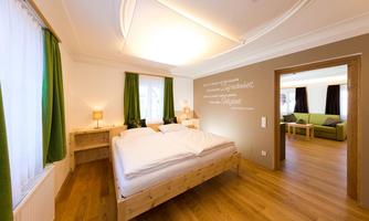 A modern bedroom with light wood and green curtains. In the background, an adjacent living room with a sofa can be seen.