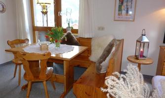 A cozy dining area with a wooden table and chairs. The window offers a view of nature, and there are decorative elements like flowers and a lantern.
