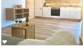 A modern kitchen with wooden cabinets and an open living area. A small table with two wine glasses and a bottle of wine complements the cozy atmosphere.