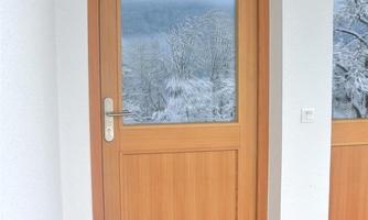 A wooden door with a glass window behind which a winter landscape is visible. In front of the door lies a doormat with various designs.