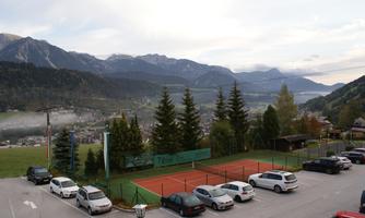 A tranquil landscape with a view of mountains and a tennis court. In the foreground, several cars are parked in a parking lot.