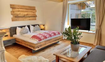 A cozy bedroom with a wooden bed and red bedding. The room has large windows and a modern TV corner.