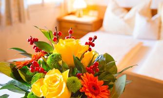 A colorful bouquet with yellow roses, red berries, and green flowers. In the background, a cozy bedroom can be seen.