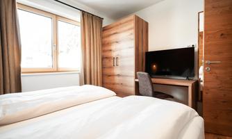 A modern bedroom with two beds and a desk. The room has large windows and a television.
