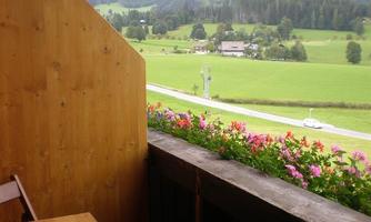 A balcony with a wooden table and colorful flowers. In the background, the impressive mountains and a green landscape can be seen.