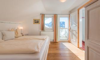 A cozy bedroom with two beds and a wooden floor. Through the windows, you can see the mountains and the entrance to the balcony.