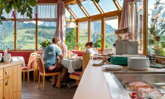 A cozy restaurant with a glass front and a magnificent view of the mountains. Two guests enjoy their meal at a table, surrounded by a warm, inviting atmosphere.