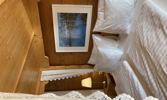 A cozy bedroom with wooden walls and two white pillows. A picture of a landscape and a lake hangs on the wall.