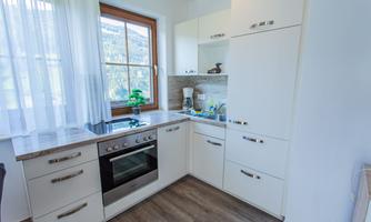 A modern kitchen with white cabinets and an oven. Bright light comes through the window, and there is a small plant on the countertop.