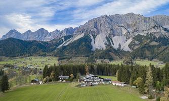 A picturesque landscape with high mountains in the background. In the foreground, there are green meadows and several buildings.