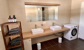 A modern bathroom with two sinks and a large mirror. On the right side, there is a washing machine and towels are neatly stacked.