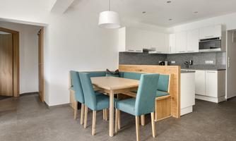 A modern dining room with a wooden dining table and turquoise chairs. Nearby is a simple, functional kitchen.