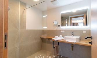 A modern bathroom with tiled walls and a large mirror. On the wooden shelf, towels and toiletries are neatly placed.