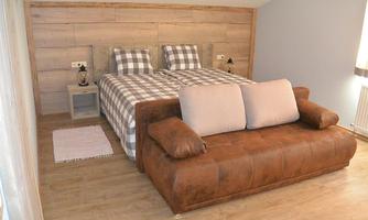A modern bedroom with a double bed and beige pillows. The room has wooden walls and a cozy leather sofa.