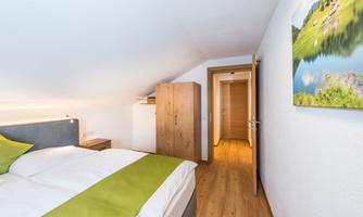 A modern bedroom with a large bed and a green duvet. The room features bright wood and access to another room.