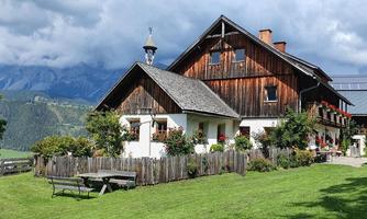 A rustic farmhouse with a wooden facade and blooming windows. In the foreground, green grass and a beautiful view of the mountains.