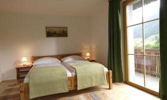 A cozy bedroom with two beds and soft green blankets. A large window lets in plenty of daylight and offers a view of the countryside.
