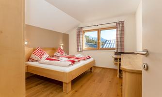 A cozy bedroom with a double bed and traditional red pillows. The room has a window overlooking the landscape and warm wooden floors.