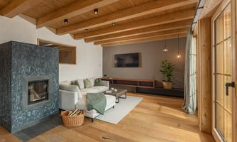 A modern living room with wooden ceiling beams and a cozy sofa. A fireplace and large windows create a bright and inviting atmosphere.