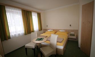 A bright bedroom with a double bed and yellow bedspreads. There is a small table with two chairs and large windows with green curtains.