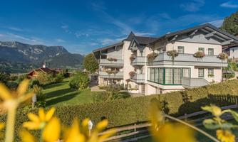 A cozy building with a balcony and floral decorations, surrounded by green meadows. In the background, gentle mountains and a blue sky can be seen.