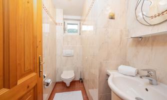 A modern bathroom with white tiles and a wooden frame. There is a toilet, a sink, and a window that provides natural light.