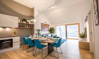 A modern kitchen with a dining area and blue chairs. Plenty of daylight streams through large windows, creating a bright, inviting atmosphere.