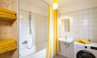 A modern bathroom with a bathtub, shower, and sink. It contains yellow towels and a washing machine.