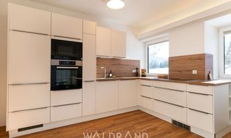 A modern kitchen with white cabinets and wooden accents. It features a cooktop, an oven, and large windows that let in plenty of daylight.