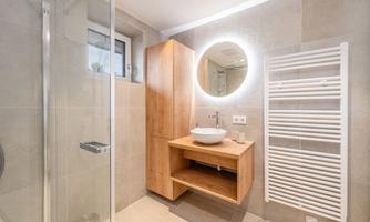 A modern bathroom with a shower, a wooden vanity, and a round mirror with light. The walls are in light beige and gray tones.