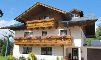 A beautiful house in Alpine style with a wooden balcony and colorful flowers. In the background, ski lifts and a green landscape can be seen.