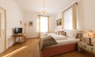 A bright bedroom with a double bed and wooden floor. There is a television, a table, and decorative lamps.