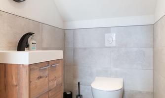 A modern bathroom with a simple wooden dresser and a wall-mounted toilet. The walls are painted in neutral shades of gray, and a round mirror complements the decor.