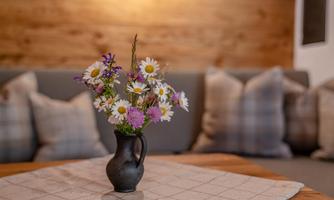 A beautiful bouquet of flowers in a black vase is on a wooden table. Cozy pillows can be seen in the background.