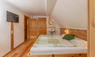 A cozy bedroom with two beds, wooden floors, and a TV wall. The walls are light, and a small washbasin is also available.