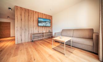 A modern living room with a comfortable sofa and a small table. A television is mounted on the wall and there is a warm wooden floor.