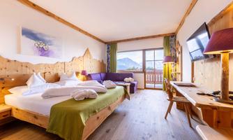 A cozy hotel room with wooden paneling and large windows. It offers a view of the mountains and is equipped with a double bed, a seating area, and a desk.