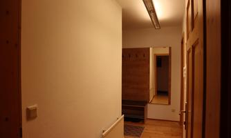 A narrow hallway with white walls and wooden flooring. On the right side, there is a bay window with a bench and coat rack.