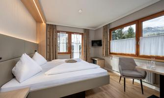 A modern bedroom with a double bed and large windows. The room is bright and friendly with a desk and a television.