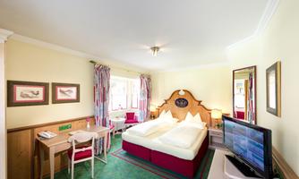 A cozy hotel room with a double bed and a television. The walls are painted in a light shade of yellow, and there is a table with chairs.