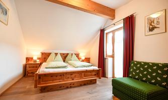 A bright bedroom with two beds and wooden furniture. A cozy armchair and large windows provide natural lighting.