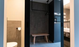 A modern bathroom with a toilet and a wooden bench. The walls are in neutral tones and the atmosphere is minimalist.