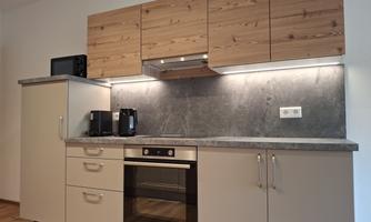 A modern kitchen with wooden cabinets and a gray countertop. The equipment includes a refrigerator, an oven, and a coffee machine.