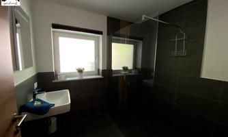 A modern bathroom with a shower and a sink. It has large windows and is done in neutral colors.
