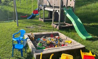 A playground with a slide, swings, and a sandbox. Colorful toys and chairs are scattered around the sandbox.