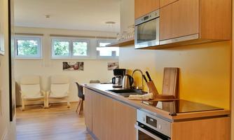 A modern kitchen with wooden cabinets, a cooktop, and a kettle. In the background, there are cozy seating areas with bright chairs.