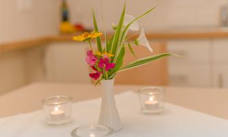 A simple table with a white vase containing colorful flowers. Around the vase, there are candle holders with burning tea lights placed.