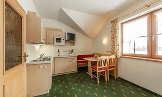 A modern kitchen with wooden cabinets and a dining area. Floral patterns are visible on the carpet and light streams through the windows.