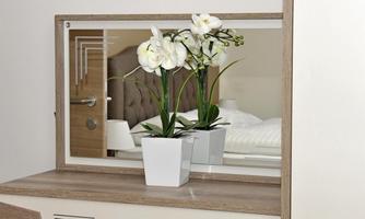 A modern mirror with a white orchid in a pot stands on a dresser. In the background, a cozy bedroom is visible.